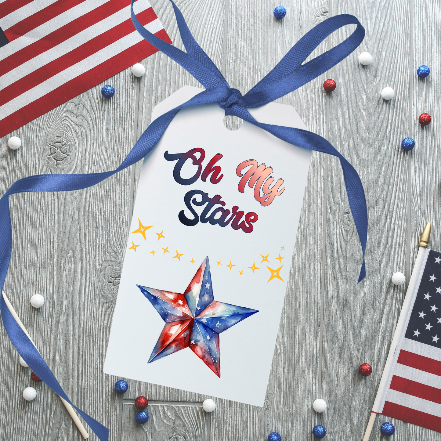 Decorative tag with 'Oh My Stars' text and star design, tied with a blue ribbon on a wooden surface with American flags and stars.